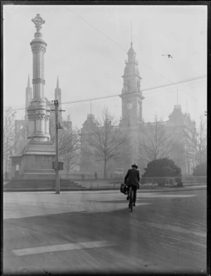 The Rev Thomas Burns Monument within the Octagon, Dunedin, with man on bicycle foreground and Saint Paul's Cathedral and Town Hall behind