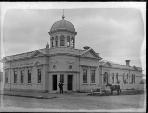 Bank of New Zealand building, Pahiatua, Tararua district, with a man standing out front, and a horse tied up at railing on street verge