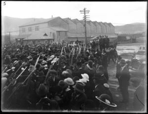 World War I troops on parade with rifles and bayonets, with crowd of people looking on, with large dockside building beyond, Wellington wharf area