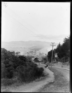 Looking north to Dunedin's Saint Clair Beach, down Bedford Street with adult and two children walking up next to bush covered hillside, to residential housing, Victoria Road and racecourse beyond