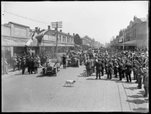 Crowd of people lining the streets and other vantage points, with other military personnel, to welcome home a procession of invalided World War I soldiers transported within cars, brass band foreground getting ready to play, Christchurch City