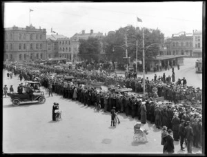 Crowd of people in two lines welcoming home invalided World War I soldiers transported within cars, trams and Reuters Telegram Company building Cathedral Square beyond, Christchurch City