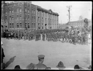View of World War I soldiers on parade with crowds of people looking on, in front of large brick building, with dock area warehouses beyond [possibly Wellington harbour?]
