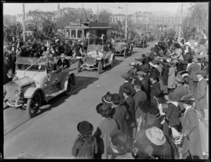View of a procession of cars carrying dignified looking people, some wearing medals, crowd looking on, horse drawn coaches with commercial buildings beyond
