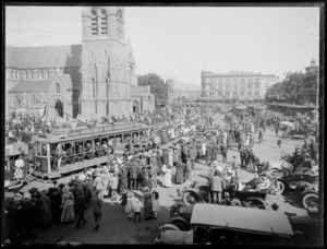 View of a procession of World War I soldiers around Christchurch Cathedral with army band on truck, followed by cars with troops, crowds of people looking on from trams, cars or on the street