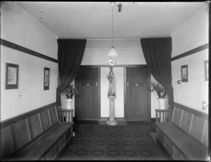 The hallway scene of [Hotel Ambassadors, Manchester Street, Christchurch?], showing rooms 31 and 32, with plants on plant stands, a lady lamp, wooden seats and prints on the walls