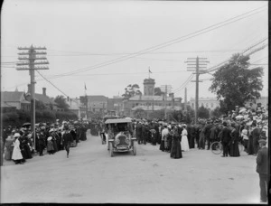 Crowds gather to watch motor cars in the parade, on an unidentified street, Timaru, with the clock tower in the background, to celebrate the coronation of George V