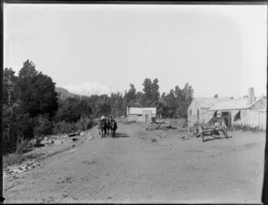 Waimarino blacksmith's shop, Taumarunui County, with two men on a cart and three horses, and a horse standing next to a shed alongside a cart