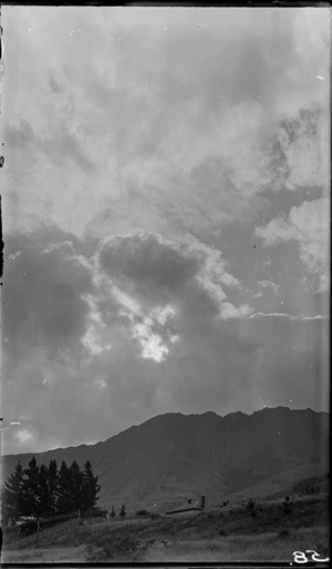 View of a grassy slope from below, including the roof of a house, and clouds over a mountain in the distance, Queenstown