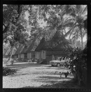 A scene in Korolevu, Fiji, showing a row of huts with thatched roofs, with an Austin 1100 car and an unidentified woman standing alongside