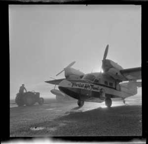 Tourist Air Travel aeroplane, on tarmac [at an aerodrome?], Invercargill, including an unidentified man on a tractor in background
