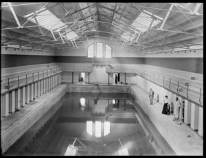 Interior of the Christchurch Municipal Baths building, with a woman and child and three men standing alongside the pool