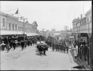 Crowds gather to watch the parade, with a team of bullocks pulling a wagon on a street lined with commercial buildings, Timaru, to celebrate the coronation of George V