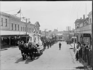 Crowds gather to watch the parade, led by a horse-drawn carriage with a group of unidentified men dressed in white robes and beards on a street lined with commercial buildings, Timaru, to celebrate the coronation of George V