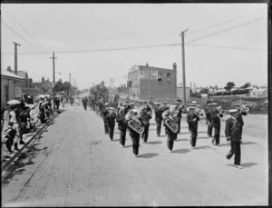 Timaru Volunteer Fire Brigade parade, led by a brass band, on an unidentified street which includes the business premises of W Healey Sanitary plumber and gasfitter, Timaru, to celebrate the coronation of George V