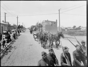 A team of horses pulling a cart with a boat full of men in the parade, led by soldiers carrying firearms, on an unidentified street which includes the business premises of W Healey Sanitary plumber and gasfitter, Timaru, to celebrate the coronation of George V