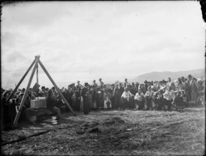 Unidentified event in a field, showing a group of Maori women performing a dance, accompanied by a violin player seated under a pulley, and observed by a crowd including mounted militia, location unidentified