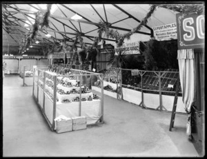 Stands with various produce on display, showing a sign for Matangi Orchards for export apples and other produce grown at the orchards, Central Otago Fruitgrowers' Association Exhibit sign, on the right side and Dawsons Ltd, jewellers, silversmiths and opticians sign in the background, possibly a trade show