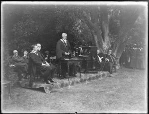 Unidentified staff members of St Andrew's College, Christchurch, showing a man standing behind a table, giving a speech