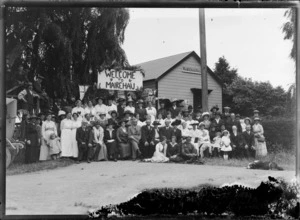 A group portrait outside the Mairehau Hall, beneath a 'Welcome To Mairehau' sign, North Canterbury