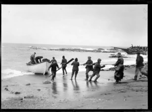 Beach scene, showing a group of men using a rope to pull a dinghy onto shore, with a shipwreck further along beach, possibly Christchurch