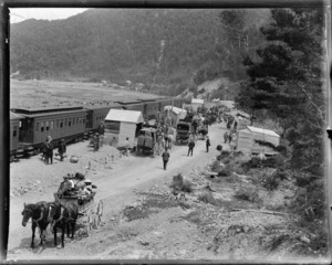 Halpin Creek railway station, showing a train stopped beside a group of corrugated iron sheds, with horse-drawn carriages on road alongside station, Selwyn District, Canterbury