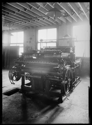 A printing press inside a brick building which has large windows and gas lamps hanging from ceiling, possibly Christchurch