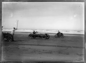 At the starting line, a man holds up the starting flag for a race between the Vauxhall racing car and the Indian racing bike on the beach