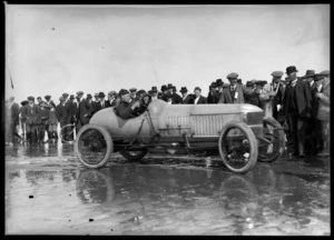 A man in a Vauxhall racing car, with crowds of people on the beach looking on