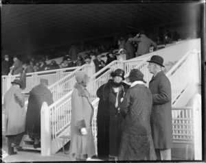 Horse racing, three women and a man standing in front of the grandstand talking