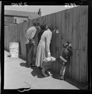 Unidentified passers-by, including a small boy with a satchel and a woman with her shopping basket, look through holes in a fence at the construction of St Paul's Cathedral, Molesworth Street, Wellington