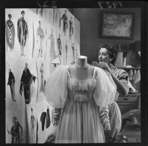 An unidentified woman making a costume for a New Zealand Players Theatre Trust production of The Merchant of Venice, including sketches by Raymond Boyce pinned to a wall alongside