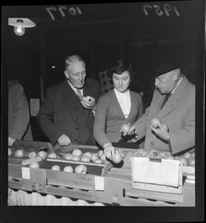 Apples on a conveyor belt are sampled by Prime Minister Walter Nash, an unidentified young woman, and elderly man, at the Apple and Pear Board's celebration of one million boxes of fruit produced, Nelson