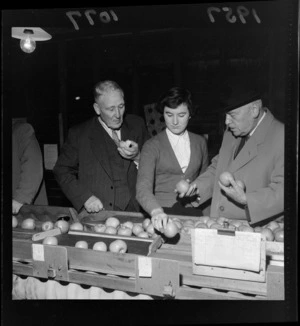 Apples on a conveyor belt are sampled by Prime Minister Walter Nash, an unidentified young female worker, and an elderly man, at the Apple and Pear Marketing Board's celebration of one million boxes of fruit produced, Nelson