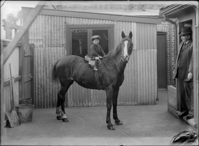 Outdoors back area of buildings, horse stables with buckets and shovels, an unidentified older man looking at young boy sitting on the back of a racing horse, probably Christchurch region