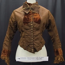 Blouse - Fitted blouse in brown brushed cotton with a polished cotton lining. Darted front to a point at waist. Inset of copper coloured velvet on upper bodice and around plain round collar, with a large bow of brown ribbon edged with a gold thread. Long two piece sleeve with a small gather at elbow for arm movement, heavy cuff is copper velvet with matching coloured machine lace frill at edge, lining of arms is a striped cotton. Opening down centre front closed with 14 buttons and button holes from waist to bust and then to neck is closed with metal hooks and cotton thread holes. Back is shaped with six panels becoming 'tails' at bottom. Label at neck reads, 'William Rattray 178 . 180 . 182 Queens Street Auckland N.Z.'.