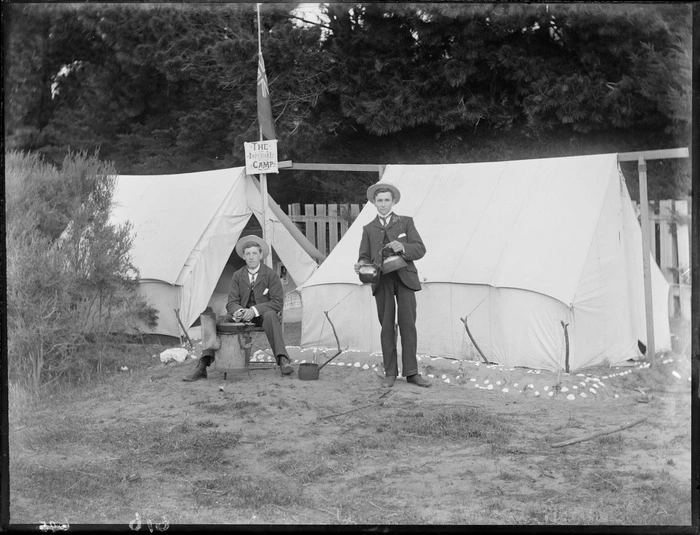 Two unidentified young men in straw hats having breakfast, one pouring tea, one cooking over a wood burner, in front of two tents with an 'The Imperial Camp' sign and flag, trees beyond, [Sumner?], Christchurch