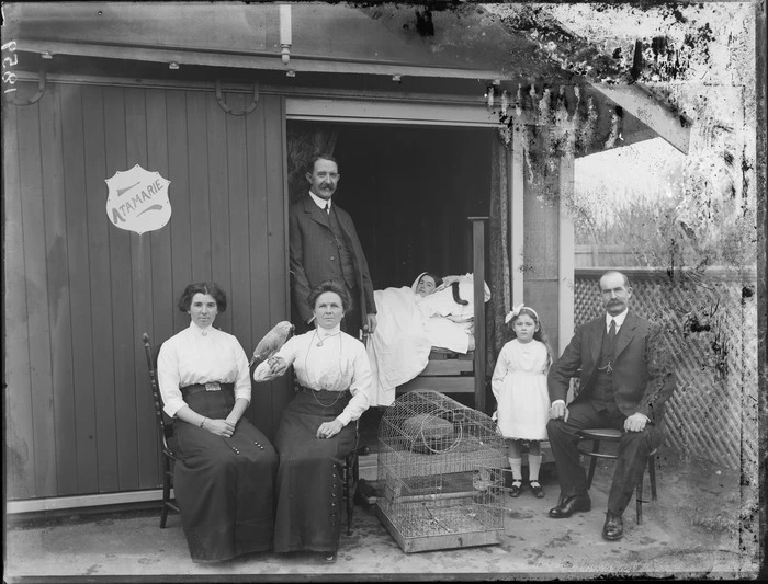 Unidentified family outside house named Atamarie with parrot and cage; a woman is lying in bed inside doorway, probably Christchurch region