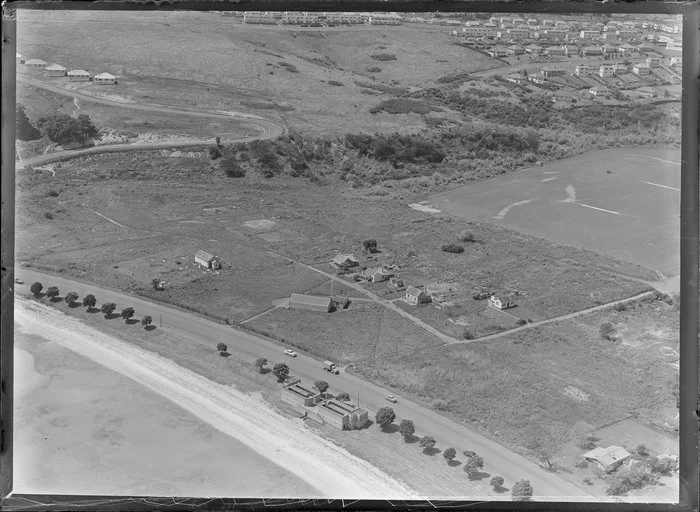 Orakei, Auckland, view of Maori settlement at Orakei Domain, Okahu Bay, with beach and Tamaki Drive with several houses and meeting house on scrubland foreground, sports field and residential houses beyond