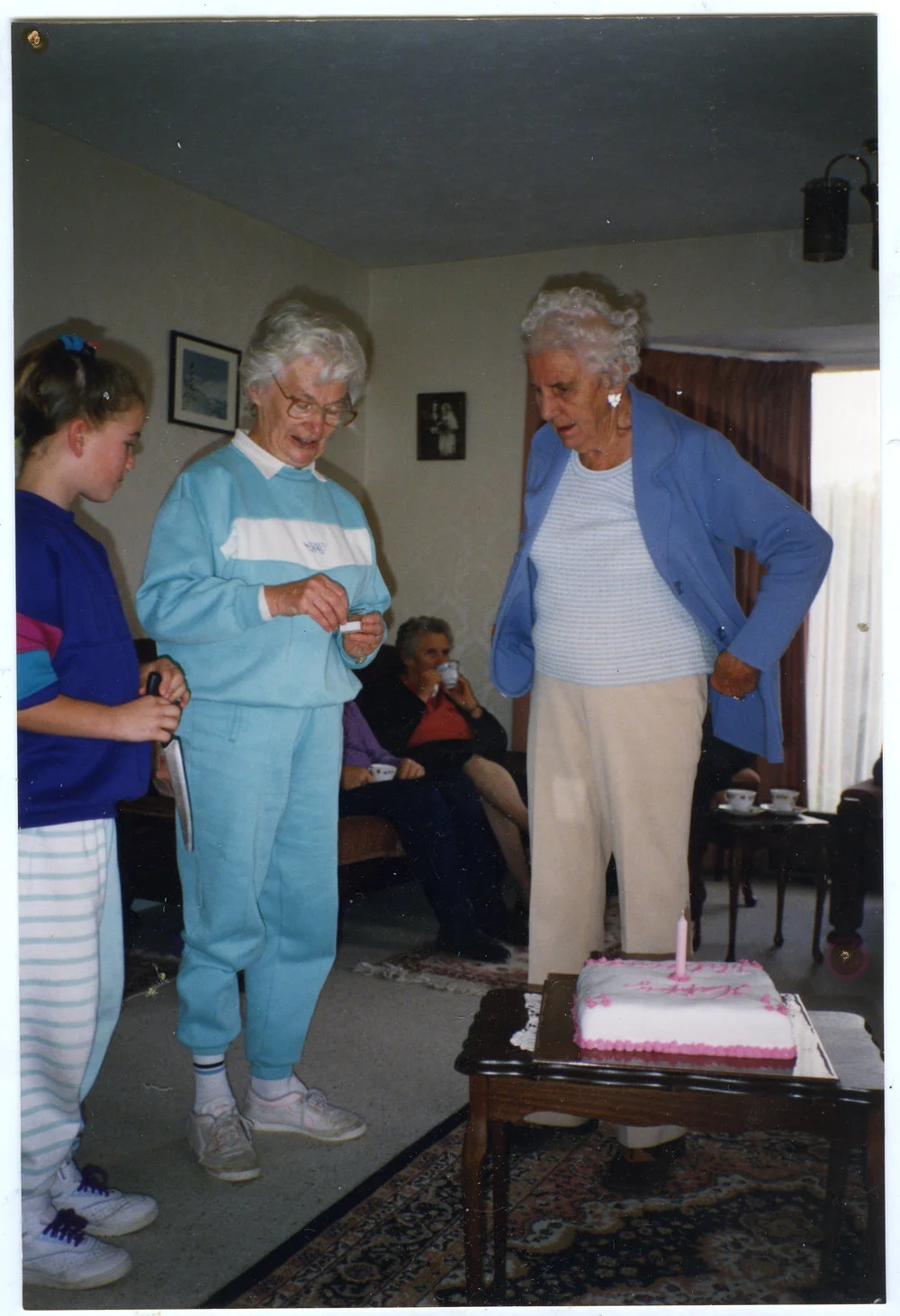 Alicia Forbes, Margaret Weir and Hilda Muff are standing in a lounge, about to light a candle on a celebration cake