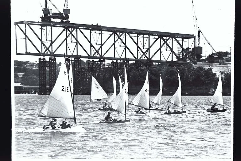 Group of children in P-class yachts sailing in front of construction work on the Auckland Harbour Bridge, 1950s