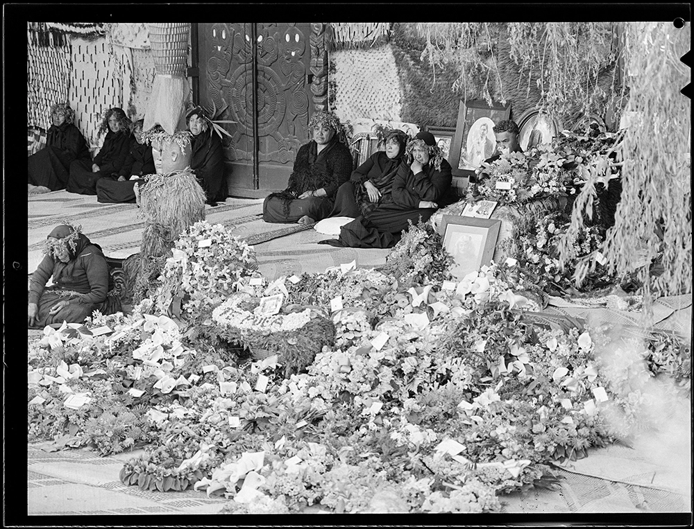 Women on whare entrance with wreaths (3) & casket