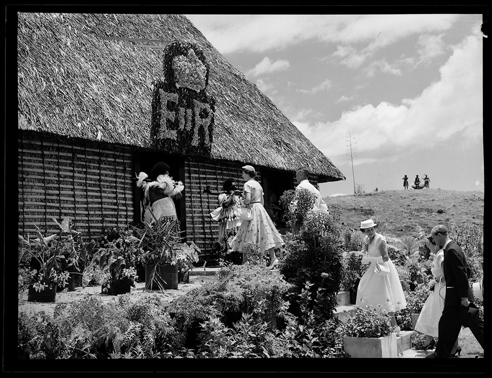 The colourful scene at Lautoka as Her Majesty was about to enter the Govenor's native style official residence. A luncheon of European and Fijian dishes was served here before the Royal visitors were entertained at a sports gathering.