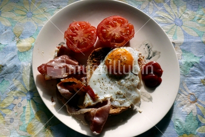 Breakfast of bacon, eggs, tomatoes and tomato sauce on retro tablecloth and plate in New Zealand.
