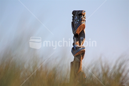 Carving of Matariki in background behind beach grass in soft focus.
