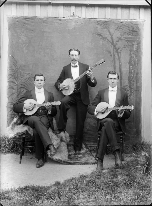 Outdoors portrait of three unidentified musicians in tuxedos, two younger men sitting with white bow ties holding mandolins, with an older man standing behind with a large moustache and black bow tie holding a banjo, probably Christchurch region