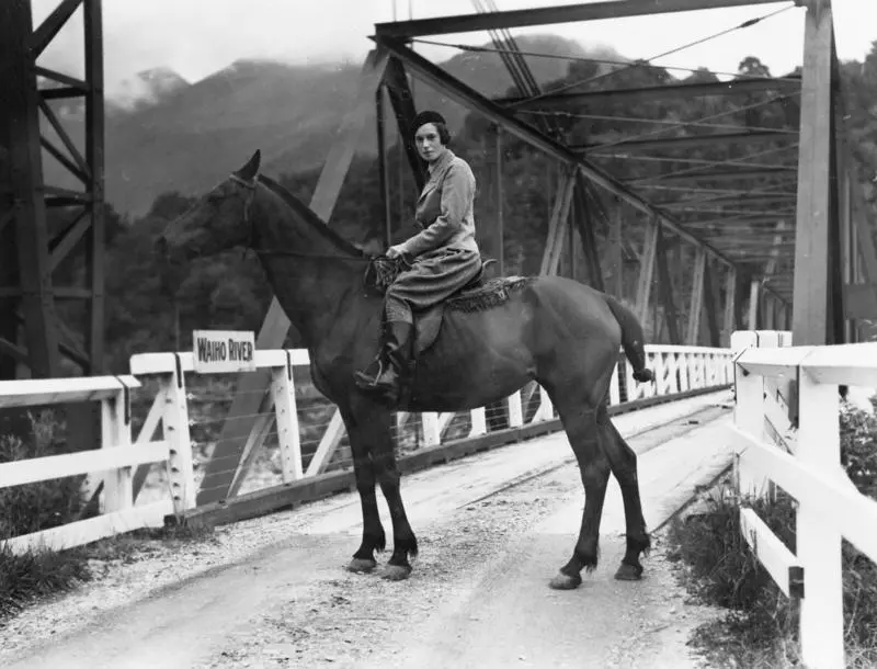 Jean Batten riding a horse over a bridge crossing Waiho River, Franz Josef / Waiau