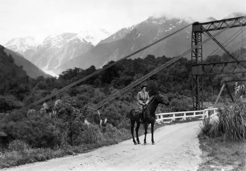 Jean Batten riding a horse approaching a bridge crossing Waiho River, Franz Josef / Waiau