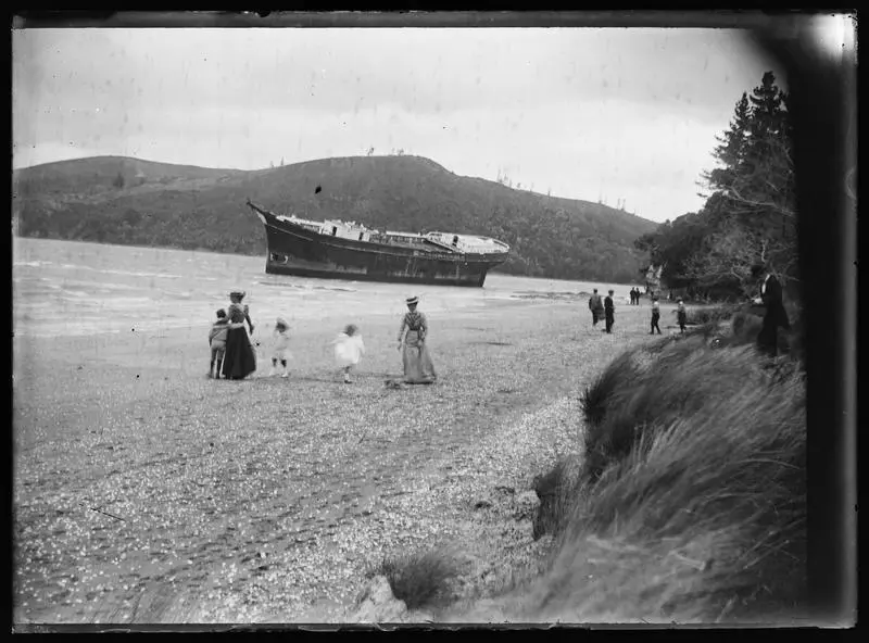 People walking on shoreline with grounded and dismasted ship in background.