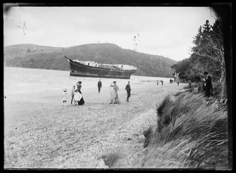 People walking on shoreline with grounded dismasted ship in background .